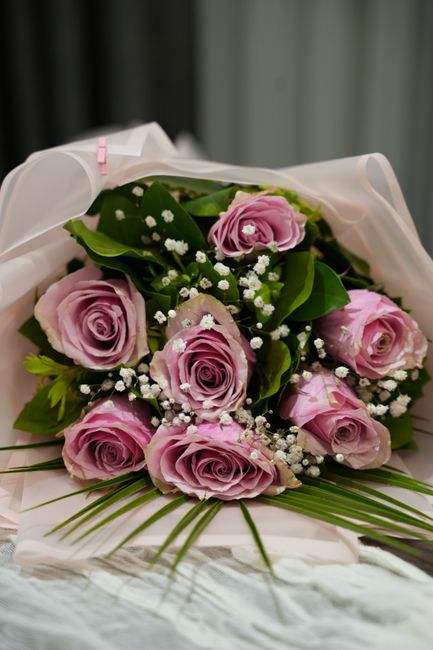 A bouquet of fresh, pale pink roses with tightly layered petals and green leaves, arranged with sprigs of delicate white baby's breath (Gypsophila) to add texture. The flowers are wrapped in white sem