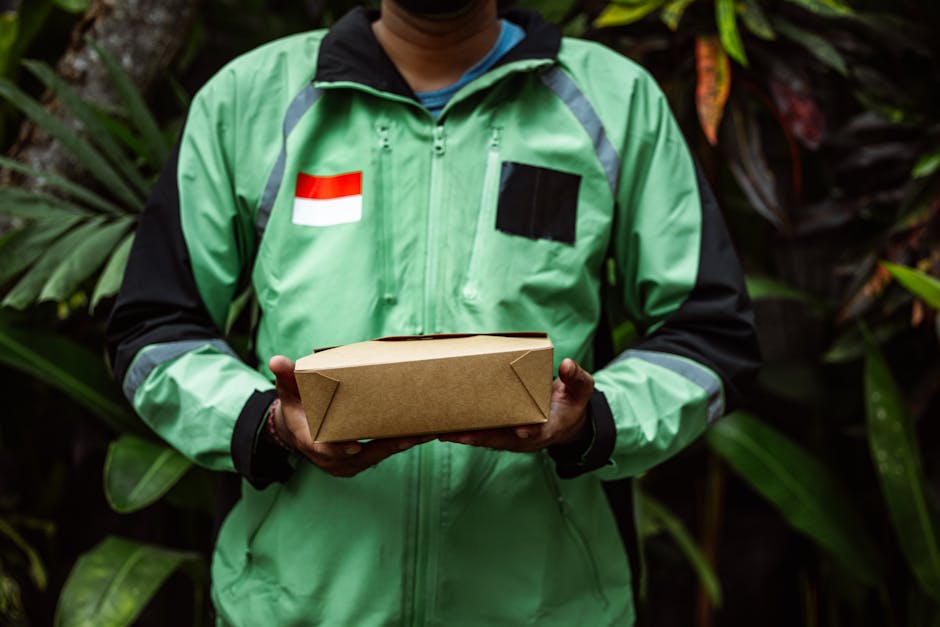 A delivery person wearing a green and black jacket with a red and white flag patch on the chest, holding a rectangular brown paper parcel in both hands. The background features lush green foliage, sug