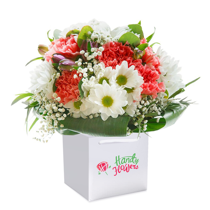 Placed inside a crisp, square white gift bag and set against a clean, white canvas, this vibrant bouquet evokes the refined comfort of a Chigwell home. The focal point is a lavish spread of coral, peach, and scarlet-red carnations, their abundant, ruffled petals capturing the eye and inviting a tactile curiosity. Amidst this sea of texture, crisp white daisy-like blooms-each with a luminous yellow-green center-shine with clarity, their upright faces imparting a feeling of cheerfulness found on sunlit mornings along Chigwell's meandering lanes. Interlaced throughout, airy white sprays of baby's breath delicately soften the overall structure, while a few closed purple-tipped buds offer an intriguing contrast and sense of evolving beauty. The outer edges are enveloped in a mantle of deep green leaves, glossy and broad, which support and define the arrangement's rounded form. Every element is meticulously arranged-each stem chosen to balance color and movement, every bloom angled to maximize freshness and appeal. Satin ribbon handles arch gracefully from the bag, suggesting easy delivery to family or friends nearby, perhaps after a visit to a Church Lane cafe or stroll in Manor Road's gardens. The branding is subtle but elegant, nodding to Chigwell's proud tradition of thoughtful gifting. Designed to suit any occasion, this bouquet stands as a testament to the artistry and neighborly warmth of Chigwell's local florists.